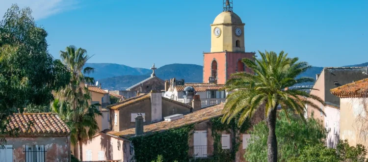 Vue sur le célèbre clocher de style baroque de l'Église Notre-Dame-de-l’Assomption à Saint-Tropez. La tour du clocher affiche ses couleurs emblématiques jaune ocre et terre de Sienne, surmontée d'une horloge et d'un campanile en fer forgé. La structure se détache sur un ciel azur lumineux, entourée par les toits de tuiles provençales du village.
