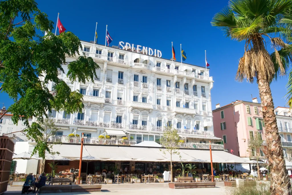 Façade ensoleillée de l'Hôtel Splendid à Cannes, un bâtiment historique du XIXe siècle de style Belle Époque avec des balcons en fer forgé. Plusieurs drapeaux internationaux flottent sur le toit à côté de l'enseigne "SPLENDID". Au rez-de-chaussée, une terrasse animée avec des parasols blancs, des tables et des chaises accueille des clients. Des palmiers et des arbres verdoyants encadrent le bâtiment sous un ciel bleu pur.