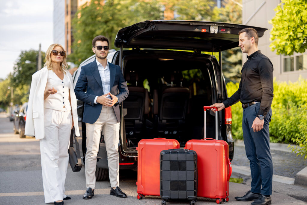 Un chauffeur privé VIP accueillant un couple d'affaires élégant à côté d'un van haut de gamme avec coffre ouvert et valises.