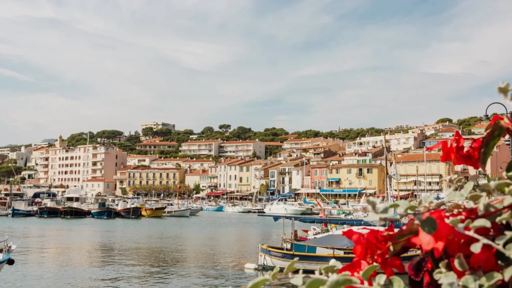 ue panoramique et ensoleillée du port de Cassis avec ses bateaux de plaisance et pointus traditionnels amarrés sur une eau paisible. En arrière-plan, les façades aux teintes pastel et ocre des maisons provençales s'alignent au pied d'une colline verdoyante. Au premier plan à droite, des fleurs rouges éclatantes apportent une touche romantique et encadrent le paysage.