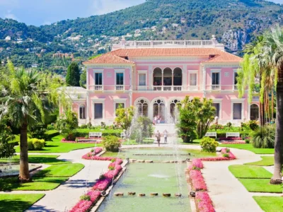 Une vue sur la magnifique façade rose d'inspiration Renaissance italienne lors d'une visite de la Villa Ephrussi de Rothschild. Le palais s'élève derrière un grand bassin d'eau orné de jets d'eau symétriques et de nénuphars, entouré par la végétation luxuriante du jardin à la française composé de pelouses tondues, de buis taillés et de hauts palmiers sous un ciel bleu parsemé de légers nuages.