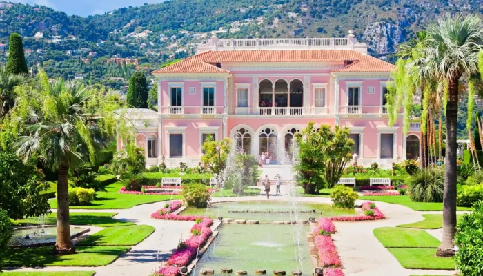 Une vue sur la magnifique façade rose d'inspiration Renaissance italienne lors d'une visite de la Villa Ephrussi de Rothschild. Le palais s'élève derrière un grand bassin d'eau orné de jets d'eau symétriques et de nénuphars, entouré par la végétation luxuriante du jardin à la française composé de pelouses tondues, de buis taillés et de hauts palmiers sous un ciel bleu parsemé de légers nuages.