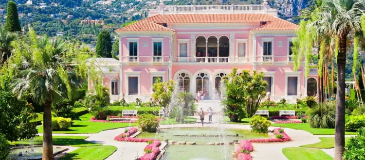 Une vue sur la magnifique façade rose d'inspiration Renaissance italienne lors d'une visite de la Villa Ephrussi de Rothschild. Le palais s'élève derrière un grand bassin d'eau orné de jets d'eau symétriques et de nénuphars, entouré par la végétation luxuriante du jardin à la française composé de pelouses tondues, de buis taillés et de hauts palmiers sous un ciel bleu parsemé de légers nuages.
