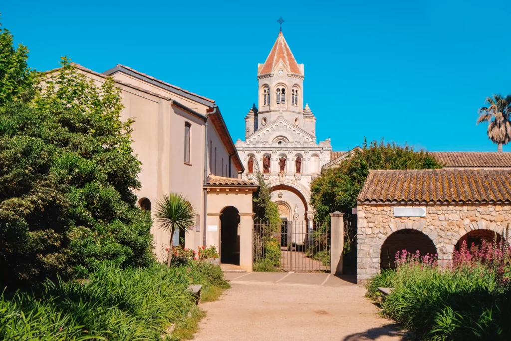 Vue ensoleillée de l'Abbaye de Lérins sur l'île Saint-Honorat. Au centre, l'église abbatiale en pierre claire se dresse avec sa tour-clocher carrée surmontée d'une flèche pyramidale rouge. La façade présente des arcatures et des statues de saints nichées. Un chemin de terre mène à une grille en fer forgé. Sur la droite, un bâtiment bas en pierre avec des arcades abrite des fleurs violettes. Une végétation méditerranéenne luxuriante, incluant un palmier et des buissons denses, encadre la scène sous un ciel bleu pur et sans nuages.