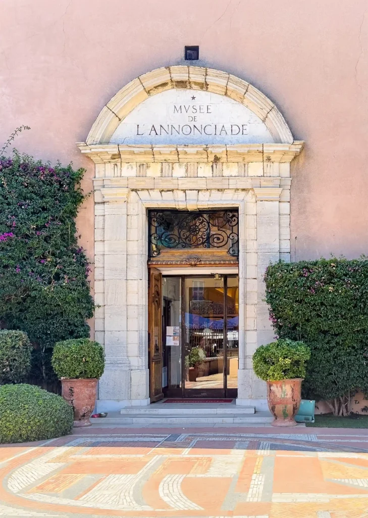 Gros plan vertical sur le portail d'entrée en pierre sculptée du musée de l'annonciade saint tropez. Le porche monumental en arche de pierre blanche surmonte une double porte en bois ouverte, révélant un hall d'entrée vitré avec une imposte en fer forgé noir ouvragé. L'inscription gravée « MVSEE DE L'ANNONCIADE » surmonte le linteau. De part et d'autre de l'entrée, deux grands pots en terre cuite abritent des arbustes taillés en boule, flanqués de buissons verdoyants contre le mur ocre rosé. Au premier plan, les motifs du sol pavé s'affichent sous une lumière ensoleillée.