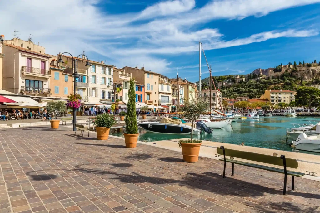 Vue ensoleillée de la promenade pavée bordant le port de Cassis, animée par des terrasses de restaurants et des passants. À droite, des voiliers et bateaux à moteur sont amarrés sur une eau calme et claire. En arrière-plan, les façades provençales colorées s'alignent sous un ciel bleu parsemé de légers nuages blancs, avec une colline verdoyante s'élevant au loin.