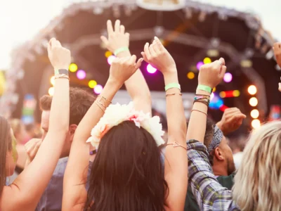 ue de dos d'un groupe de jeunes festivaliers levant les bras en l'air et portant des bracelets d'accès, face à une grande scène de concert en plein air lors d'un festival sur la Côte d'Azur. On aperçoit une jeune femme avec une couronne de fleurs dans les cheveux au premier plan sous une lumière estivale ensoleillée.