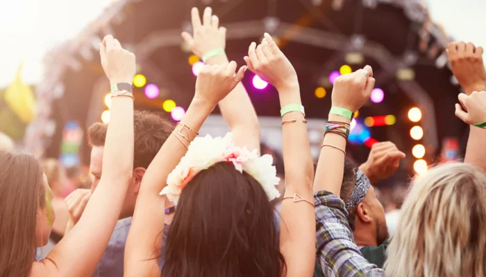 ue de dos d'un groupe de jeunes festivaliers levant les bras en l'air et portant des bracelets d'accès, face à une grande scène de concert en plein air lors d'un festival sur la Côte d'Azur. On aperçoit une jeune femme avec une couronne de fleurs dans les cheveux au premier plan sous une lumière estivale ensoleillée.