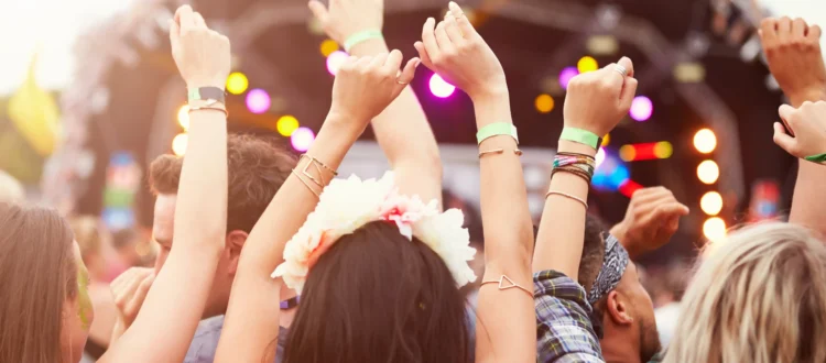 ue de dos d'un groupe de jeunes festivaliers levant les bras en l'air et portant des bracelets d'accès, face à une grande scène de concert en plein air lors d'un festival sur la Côte d'Azur. On aperçoit une jeune femme avec une couronne de fleurs dans les cheveux au premier plan sous une lumière estivale ensoleillée.
