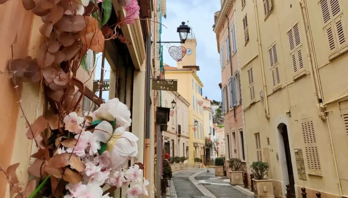 Perspective view of a narrow, cobbled alley in Vieux-Cannet, Rue Saint-Sauveur, lined with Provençal buildings with ochre and yellow facades. In the foreground on the left, a dense floral arrangement of dried brown flowers and pink and white artificial flowers (peonies and hydrangeas) frames a shop window. In the distance, aligned with the street, rises the iconic yellow bell tower of the Saint-Sauveur Chapel under an overcast sky, with a decorative heart visible near the top. @jeanpaul 2025