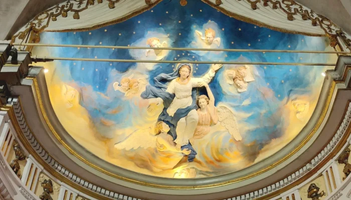 A low-angle photograph of a religious fresco adorning a semicircular dome. It depicts the Assumption of the Virgin Mary, draped in white and blue, ascending towards a deep blue, starry sky. She is surrounded by a cloud of angels and cherubs with outstretched wings, painted in shades of white, gold, and blue. The fresco is framed by a white draped curtain and an architectural frieze decorated with gilded candelabra and classical motifs. @jeanpaul 2025
