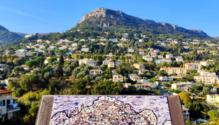 An enameled lava art interpretation plaque reproducing Jean Dubuffet's work, "View of the Sunny Side," is positioned in the foreground on a rampart. Behind the plaque, in a blurred but recognizable background, stretches the true panoramic view of the residential hills of Vence and the summit of a rocky Baou under a clear blue sky. An identification plaque at the base of the lectern bears the name "Jean DUBUFFET" and the title of the work. @jeanpaul
