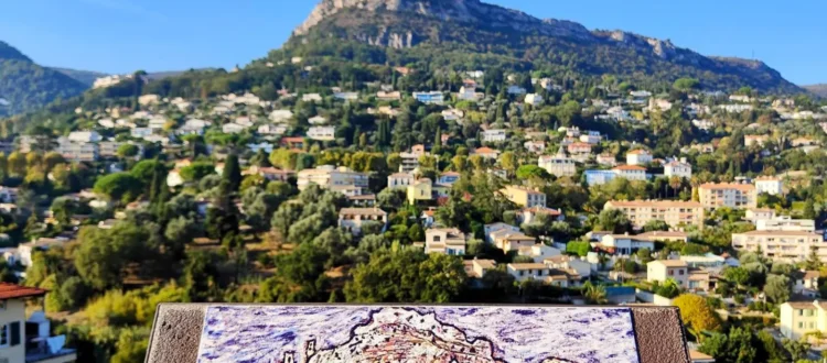 Une plaque d'interprétation artistique en lave émaillée reproduisant l'œuvre de Jean Dubuffet, « La vue sur l’Adret », positionnée au premier plan sur un rempart. Derrière la plaque, en arrière-plan flou mais reconnaissable, s'étend la véritable vue panoramique sur les collines résidentielles de Vence et le sommet d'un Baou rocheux sous un ciel bleu clair. Une plaque d'identité au pied du pupitre mentionne « Jean DUBUFFET » et le titre de l'œuvre. @jeanpaul