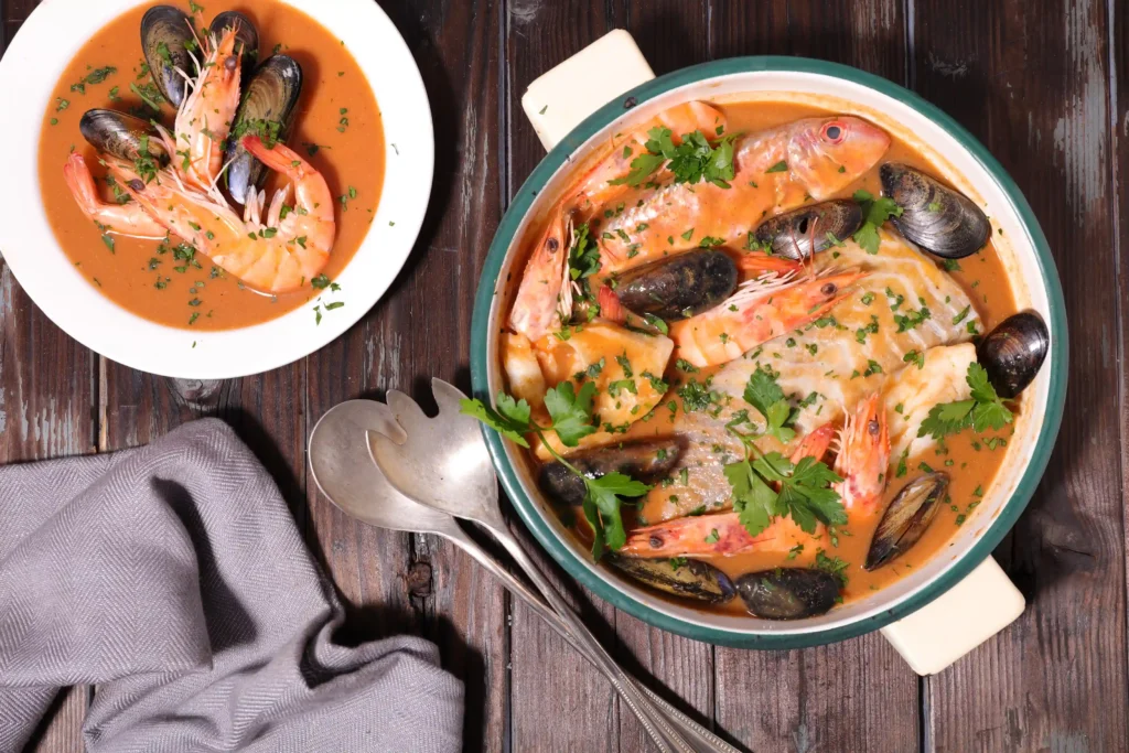Une bouillabaisse provençale traditionnelle servie dans une cocotte en céramique sur une table en bois, entourée de croûtons, d'une sauce rouille, de couverts anciens et d'un bol de service, avec des feux d'artifice en arrière-plan.
