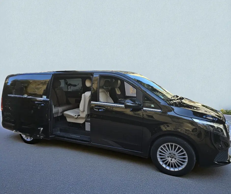 Vue latérale d'un van Mercedes Classe V XL noir brillant de la flotte EDEN CAB. La grande porte latérale coulissante est ouverte, révélant un habitacle spacieux avec des sièges en cuir de couleur crème. Le véhicule est stationné sur une route asphaltée, avec un mur texturé gris en arrière-plan.
