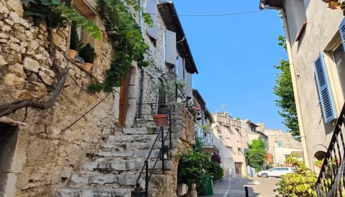 View of a picturesque medieval alley in La Colle-sur-Loup with an ancient stone staircase on the left, a climbing vine, and typical Provençal facades under a blue sky. @jeanpaul