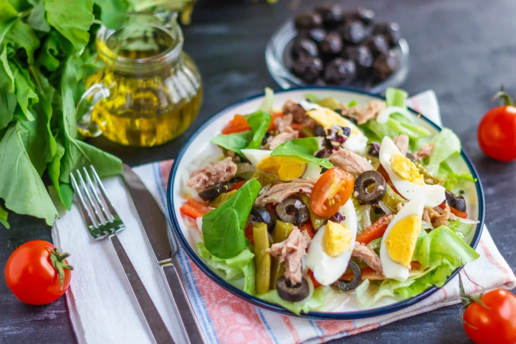 Une salade niçoise authentique présentée dans une assiette creuse, composée de thon, d'œufs durs, de tomates cerises, d'olives noires de Nice, de haricots verts et de crudités, accompagnée d'une burette d'huile d'olive.