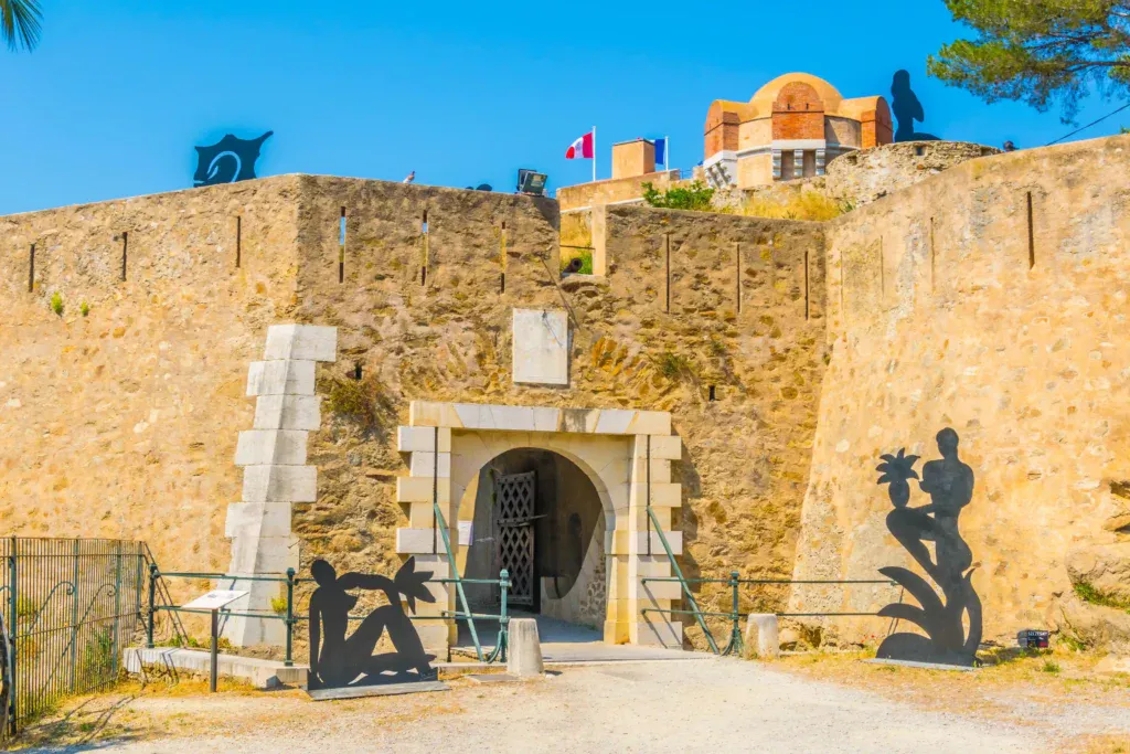 Photographie de l'entrée fortifiée de la Citadelle de Saint-Tropez. On y voit une porte d'entrée en pierre de taille blanche en arc de cercle avec un pont-levis en bois. De hauts murs défensifs en pierre ocre entourent l'entrée, surmontés d'un cadran solaire et du drapeau français. À droite, un ancien canon en bronze est exposé sur son affût en bois, et à gauche, trois oliviers sont plantés dans des pots en terre cuite.