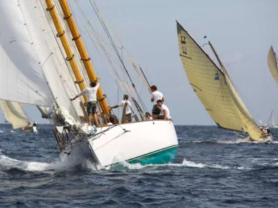 Vue rapprochée d’un grand voilier classique blanc aux mâtures en bois verni fendant les vagues en pleine mer comme lors des voiles de saint tropez. L’équipage vêtu de t-shirts blancs s’active sur le pont et au pied du mât pendant la manœuvre sous de grandes voiles blanches déployées. À l’arrière-plan, deux autres voiliers d’époque aux voiles jaunies inclinées par le vent naviguent sur une mer bleue agitation moyenne avec de l’écume blanche sous un ciel lumineux.