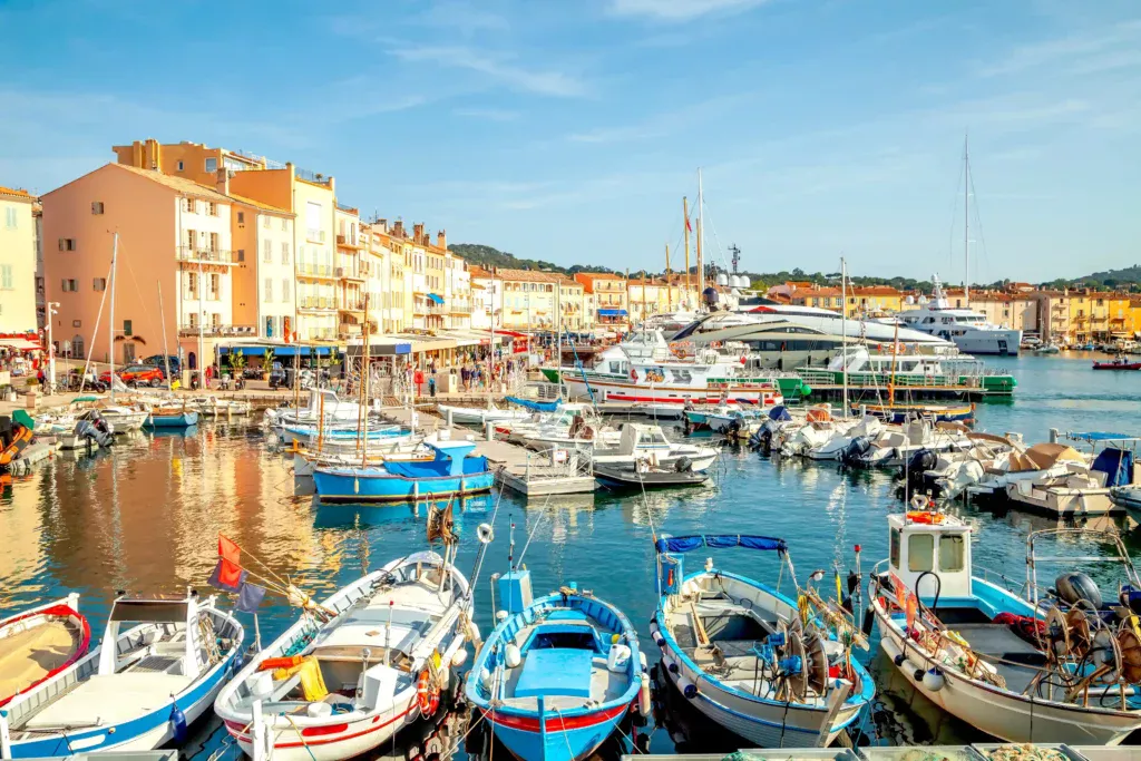 Vue sur le Vieux Port de Saint-Tropez par une journée ensoleillée. Au premier plan, plusieurs petits bateaux de pêche traditionnels et colorés, les pointus, sont amarrés côte à côte. En arrière-plan, l'eau calme du port reflète les célèbres façades ocre, pastel et dorées des bâtiments qui bordent le quai, où l'on distingue des terrasses de cafés. De grands yachts et voiliers de luxe sont ancrés plus loin sous un ciel bleu clair.